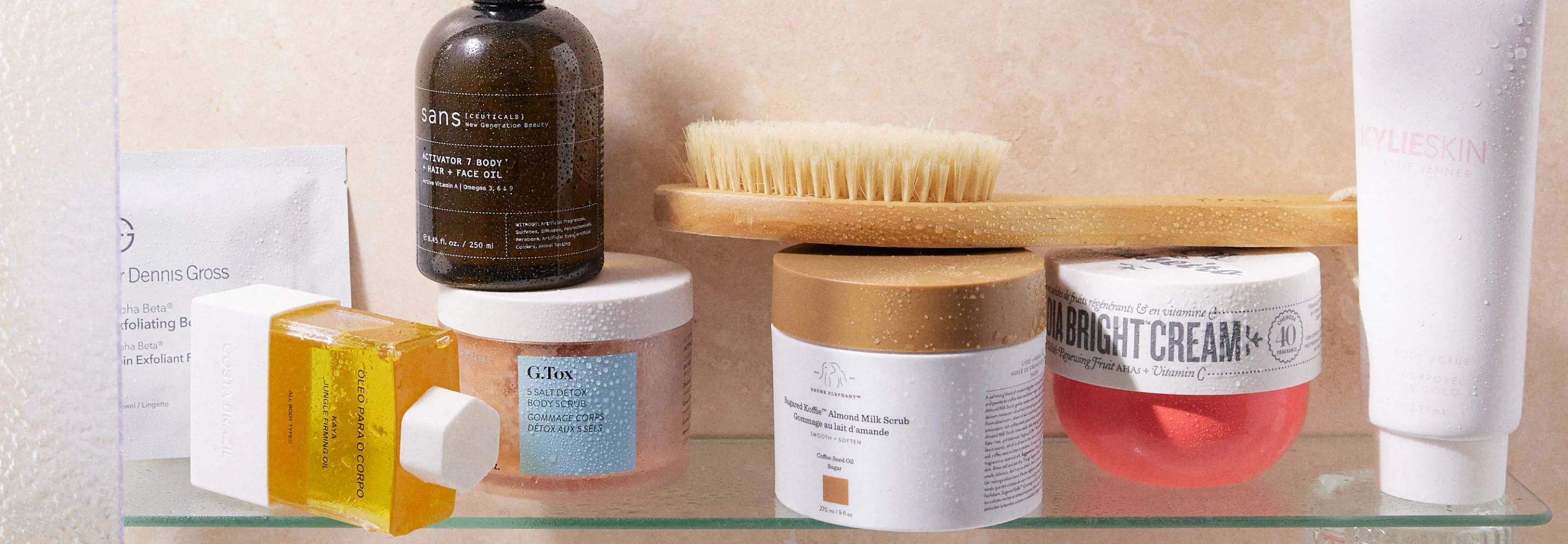 products lined up on a shower shelf: dr dennis gross alpha beta exfoliating body treatment, costa brazil firming oil, sans ceuticals activator 7 oil, goop g tox body scrub, goop dry body brush, drunk elephant koffie scrub, sol de janeiro bom dia bright cream and kylie beauty coconut body scrub