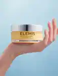 Hand holding Elemis pro-collagen cleansing balm on a blue background