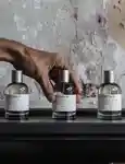3 Le Labo fragrances standing in line with a male's hand grabbing the middle one