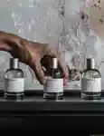 3 Le Labo fragrances standing in line with a male's hand grabbing the middle one
