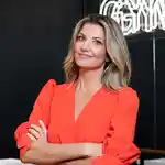 Photo of FaceGym founder Inge Theron, a blonde woman, wearing an orange blouse and standing in front of a FaceGym studio.