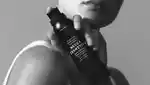 Black and white image of female model holding bottle of sunscreen mousse against her shoulder.