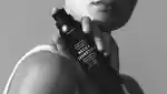 Black and white image of female model holding bottle of sunscreen mousse against her shoulder.