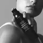 Black and white image of female model holding bottle of sunscreen mousse against her shoulder.