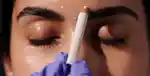 A gloved practitioner marking a client's brow area with a white pencil during a cosmetic consultation